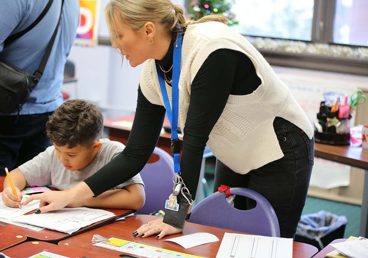 HSA Teacher smiles while kneeling beside a young student in a classroom setting.